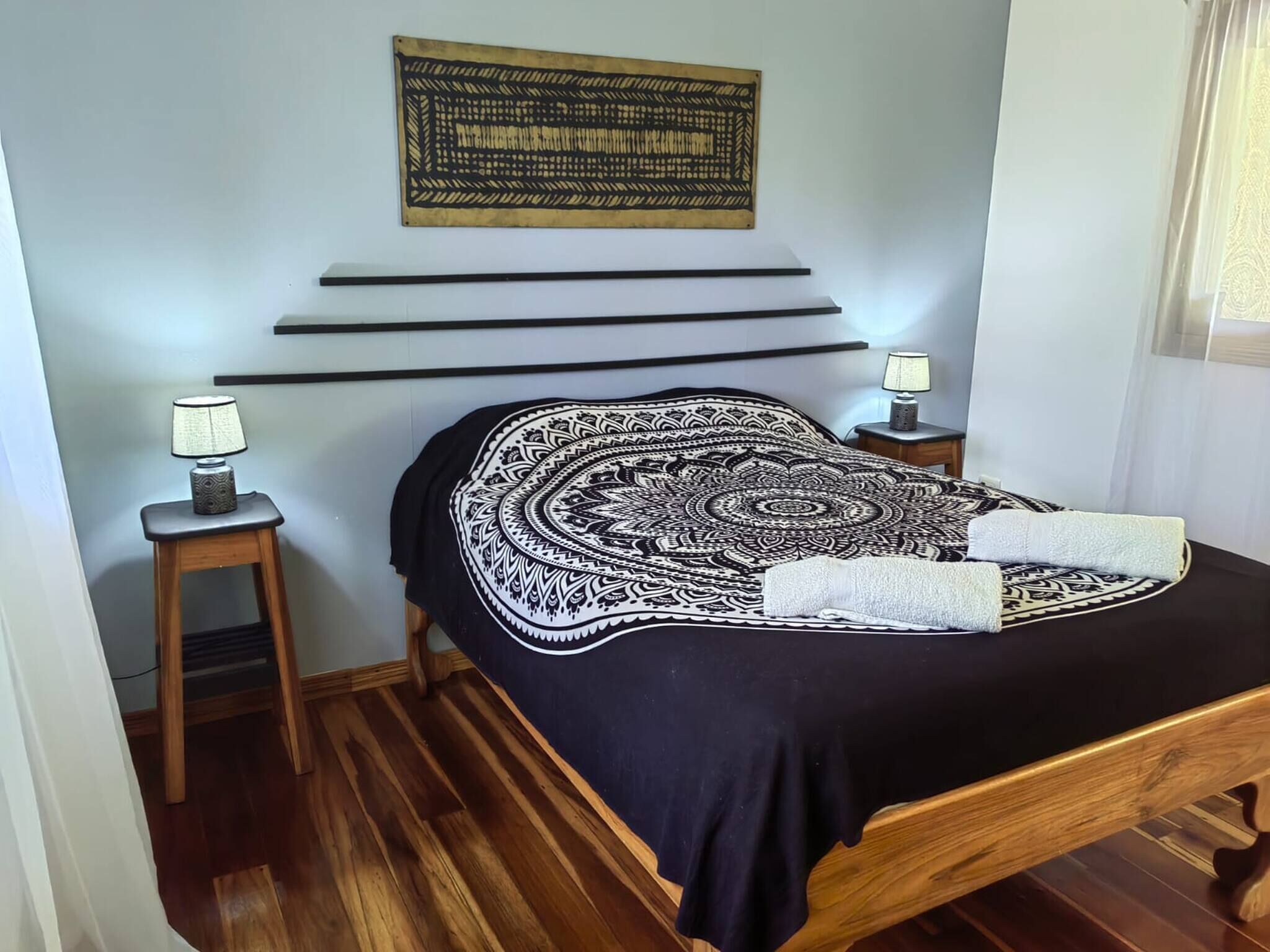 A bedroom with a bed, a wall-mounted tapestry, a bedside table with a lamp, and a wooden floor.
