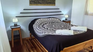 A bedroom with a bed, a wall-mounted tapestry, a bedside table with a lamp, and a wooden floor.