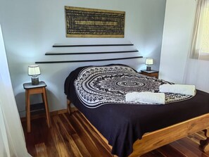 A bedroom with a bed, a wall-mounted tapestry, a bedside table with a lamp, and a wooden floor. - Brisas del Caribe # 2 (Cahuita)