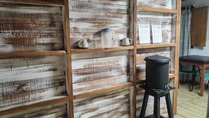Pared de madera rústica con estantes que sostienen tazas y un hervidor, un cesto de basura negro y un menú a la derecha.