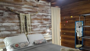 Un dormitorio con cama, almohadas, una manta, una pared de textura de madera, un macramé colgante y una estantería con objetos.