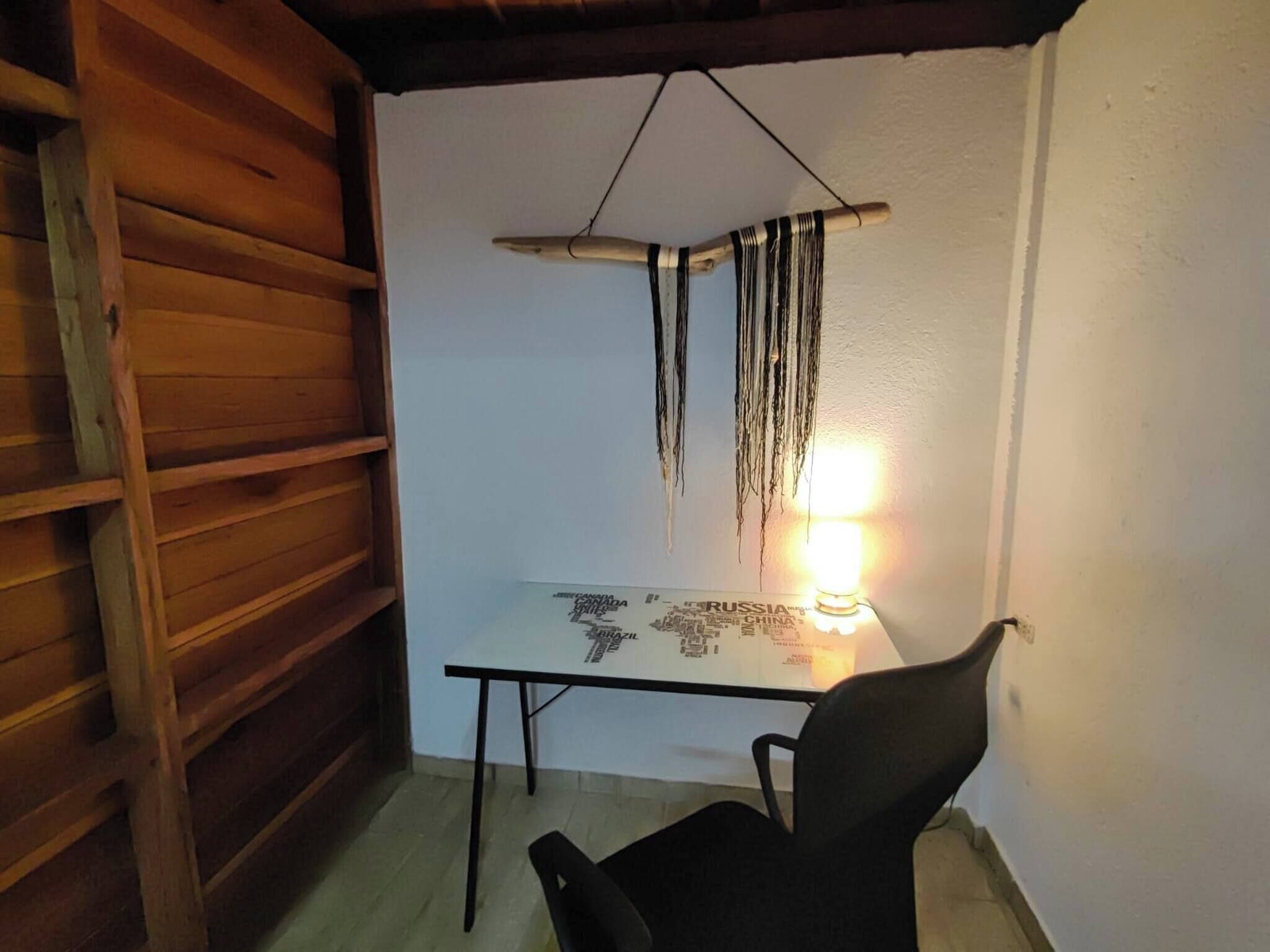 A small room with a wooden sloped ceiling, a glass table with a map, a hanging macrame decoration, and a black chair.