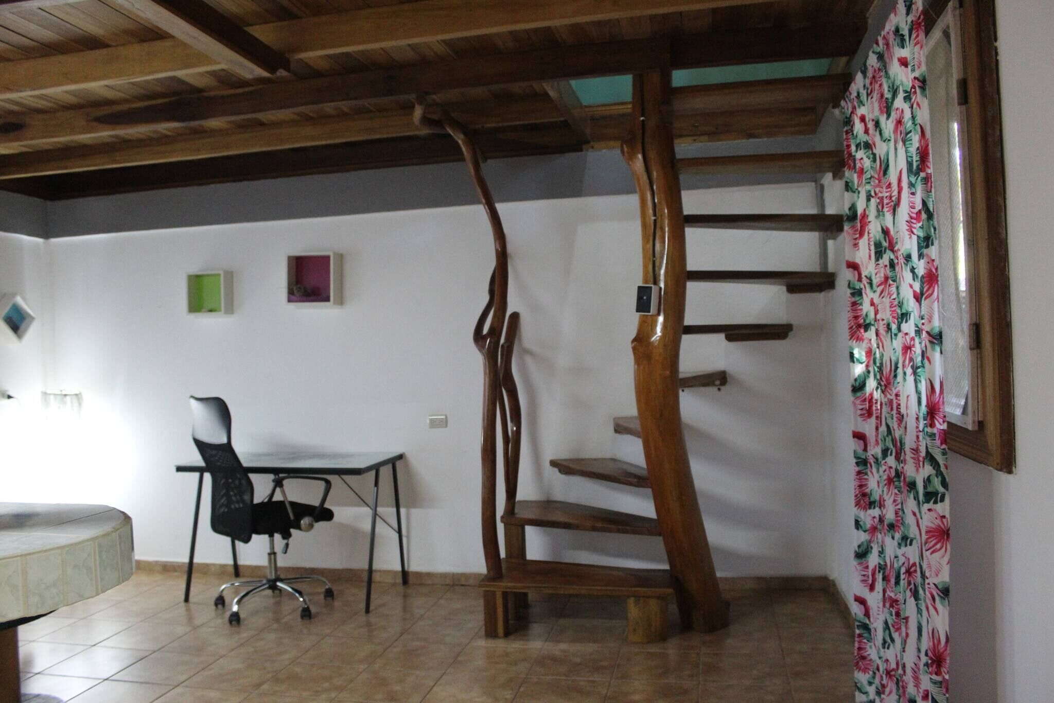 Een kamer met een houten trap, een bureau met stoel, en een raam met bloemen gordijnen.