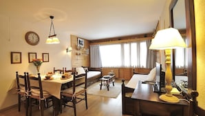 Ein gemütliches Zimmer mit Esstisch, Stühlen, einer Couch, einem Schreibtisch mit Lampe und einem großen Fenster mit Vorhängen.