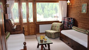 A cozy cabin room with two beds, a striped armchair, a small table with a kettle and cups, and a window with curtains. - Studio-apartment for 2-3 persons : Living room with 2 sofa-beds and 1 pull-out bed - kitchenette wit (Grimentz)