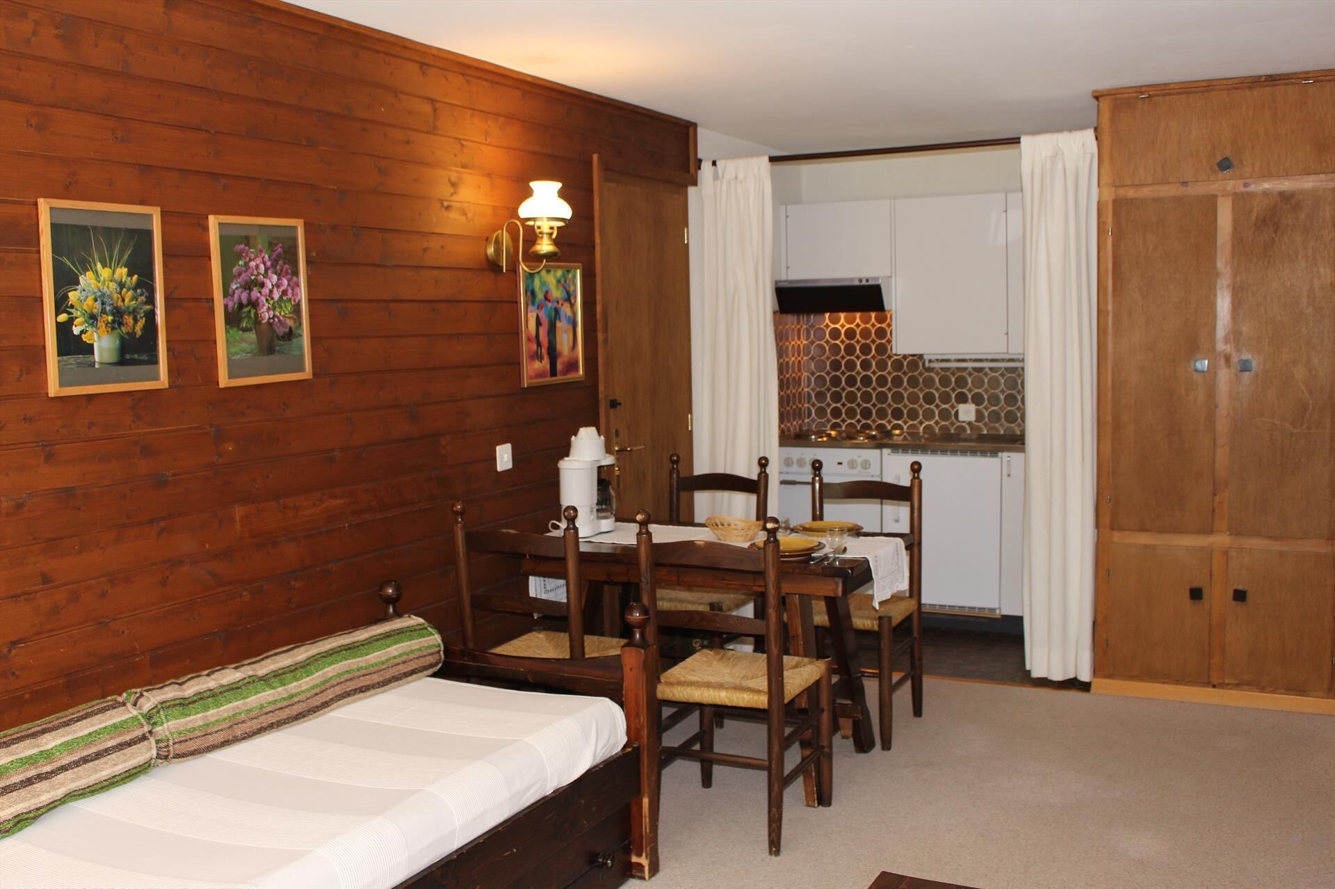 A cozy room with a wooden floor, a bed with a striped pillow, a dining table with chairs, and a kitchen area with a stove and cabinets.
