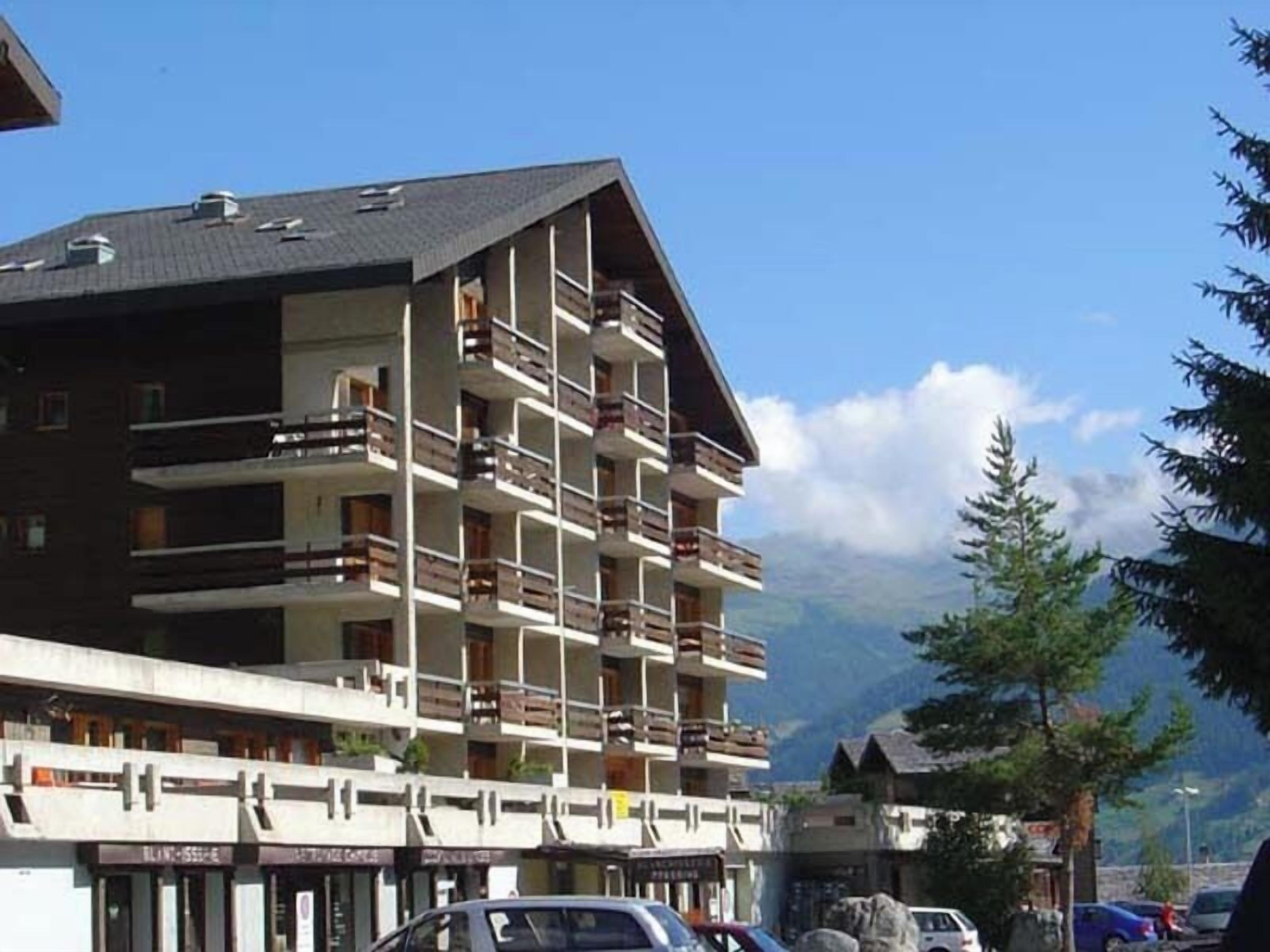 A multi-story hotel with balconies, a ski resort in the background, and a clear sky.