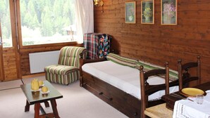 A cozy room with a wooden bed, a striped armchair, a small table with a kettle and cups, and framed pictures on the wall. - Studio-apartment for 2-3 persons : Living room with 2 sofa-beds and 1 pull-out bed - kitchenette wit (Grimentz)