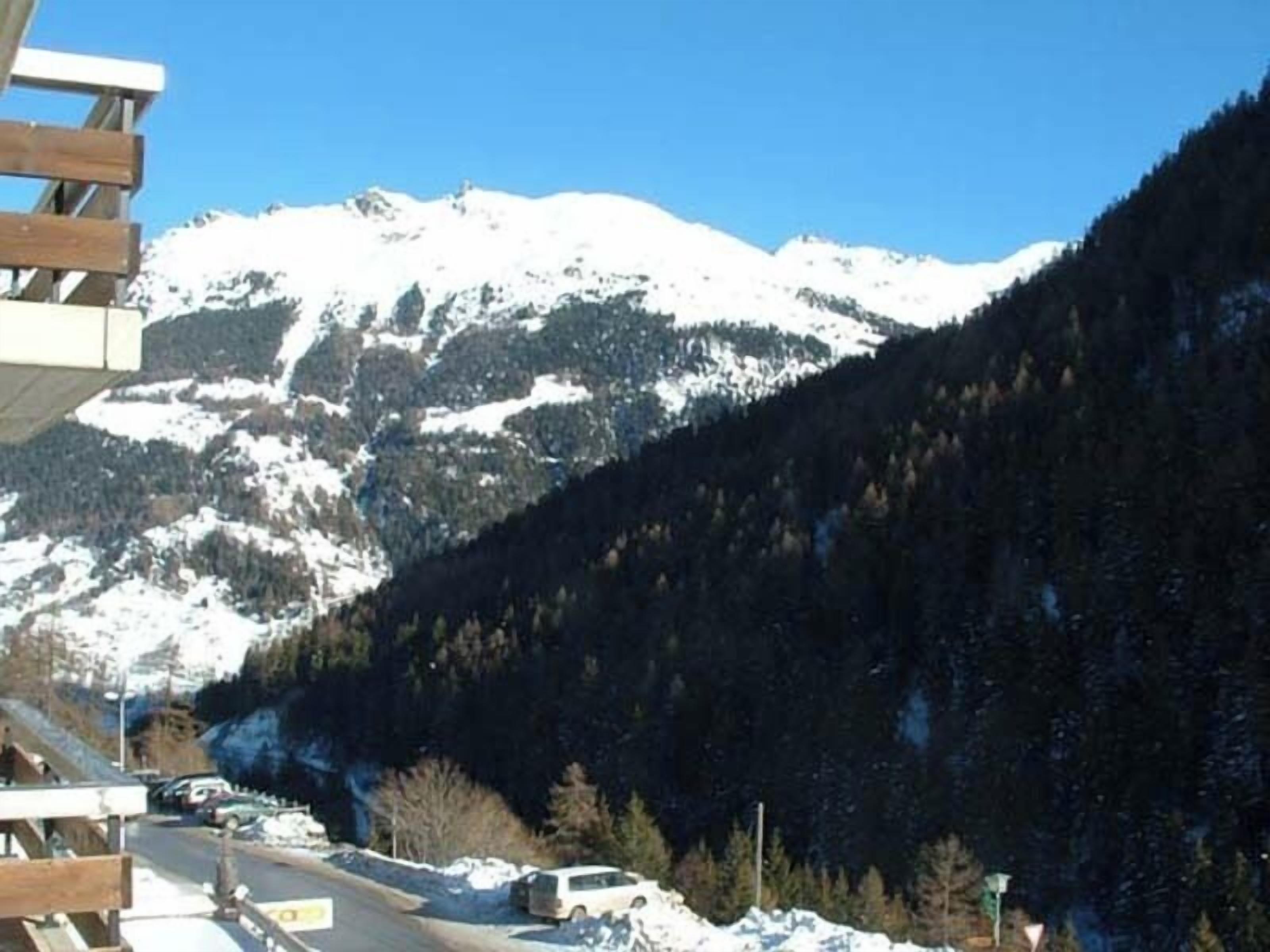 A road with a wooden bridge, snow-covered mountains, and parked cars.