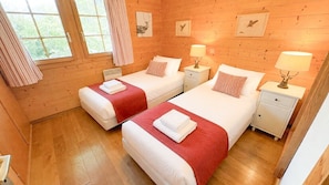 Ein schlicht eingerichtetes Schlafzimmer im Blockhausstil mit zwei Betten, jeweils mit roter Decke und weißen Kissen, sowie einem Nachttisch mit Lampe.