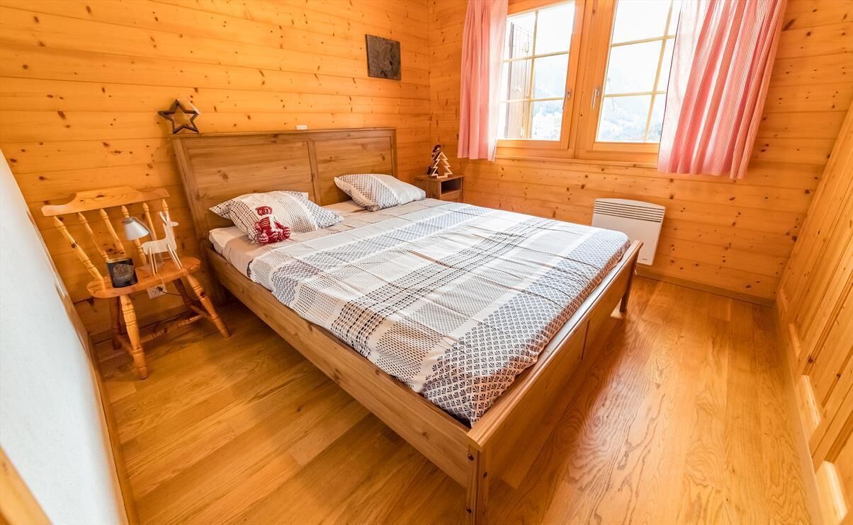 Camera da letto di una baita in legno con un letto, un comodino, una sedia e una finestra con tende.