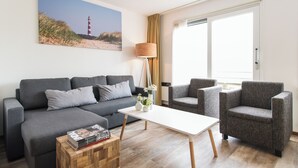 Ein modernes Wohnzimmer mit grauem Sofa, einem weißen Couchtisch und einem grauen Sessel. An der Wand hängt ein Gemälde von einem Leuchtturm, und es gibt eine Lampe sowie ein Fenster mit Vorhängen.