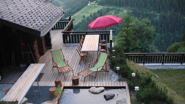 Eine Terrasse mit Gartenmöbeln und einem roten Sonnenschirm, mit Blick auf eine gebirgige Landschaft.