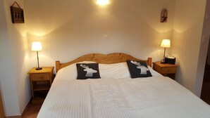 A neatly made bed with a wooden headboard, two bedside lamps, and two decorative pillows with reindeer designs. - Small, very comfortable chalet in the old village for 2 persons : living room with wood-stove - flat (Grimentz)