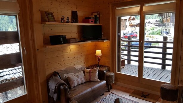 A cozy cabin interior with a wooden interior, a leather sofa, a television on a shelf, and a view of a ski resort outside. - Small, very comfortable chalet in the old village for 2 persons : living room with wood-stove - flat (Grimentz)