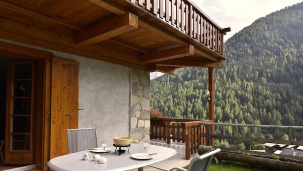 An outdoor dining area with a table and chairs, a wooden balcony, and a mountainous backdrop. - Very comfortable 4 room apartment for 6-7 persons : Living room with fireplace - kitchen with dishwa (Grimentz)