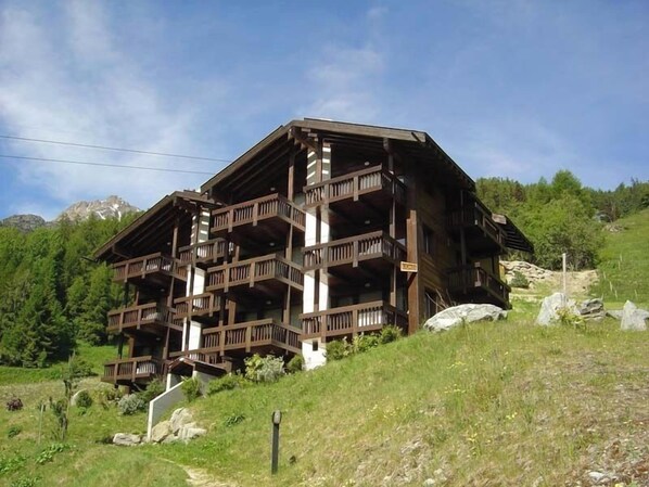 Un edificio de madera de varios pisos con balcones, situado en una colina con montañas al fondo.
