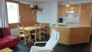 Tunnelmallinen, mökin tyylinen huone, jossa on puinen ruokapöytä, tuoleja, punainen sohva, valkoinen nojatuoli sekä pieni keittiö, jossa on liesi ja pesuallas.