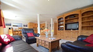 Una sala de estar con sofá, sillón y mesa de centro. Hay un televisor sobre un mueble de madera.