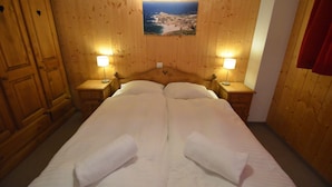 Una cama doble con sábanas blancas y dos almohadas, flanqueada por mesitas de noche de madera con lámparas, en una habitación con paneles de madera y un cuadro enmarcado de un paisaje costero en la pared.