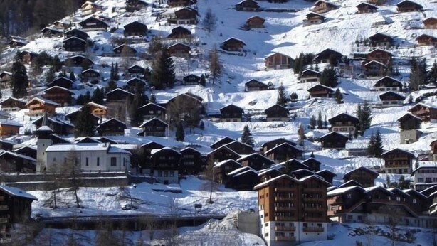 Ein verschneites Dorf mit traditionellen Holzhäusern, einige mit Balkonen, und einigen größeren Gebäuden.