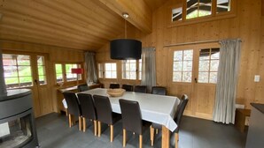 Een houten hutje met een eethoek, inclusief een tafel, stoelen, een zwarte kachel en grote ramen met gordijnen.