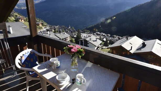 Ein Balkon mit Tisch für zwei Personen, einer Blumenvase und Blick auf ein Bergdorf.