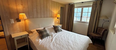 A bedroom with a wooden headboard, two bedside lamps, a bed with white bedding, a chair, and a window with curtains.