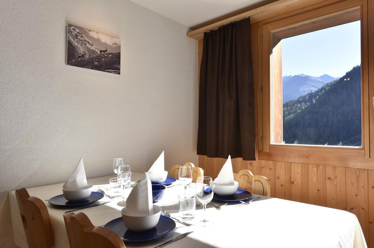 Ein Esstisch ist mit blauem und weißem Geschirr gedeckt, ein Fenster bietet Blick auf Berge, es gibt Holzvertäfelung und ein gerahmtes Bild an der Wand.