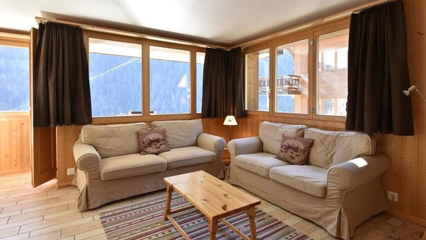 Una sala de estar acogedora con dos sofás beige, una mesa de centro de madera y ventanas grandes con cortinas.
