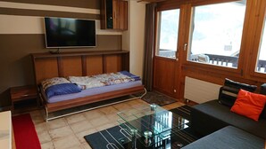A room with a bed, a TV mounted on the wall, a glass coffee table, a sofa, and a window with curtains. - 2 room apartment for 2-4 persons : Living room with sofa-bed and 1 pull-down bed - kitchen with dish (Grimentz)