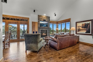 A spacious living room with a stone fireplace, leather sofas, a television, and large windows offering a view of trees.