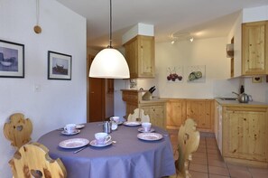 Ein Essbereich mit einem runden Tisch, gedeckt für vier Personen, umgeben von Holzstühlen. Eine große, weiße Pendelleuchte hängt über dem Tisch. Im Hintergrund ist eine Küche mit Holzschränken und einer gefliesten Rückwand zu sehen.
