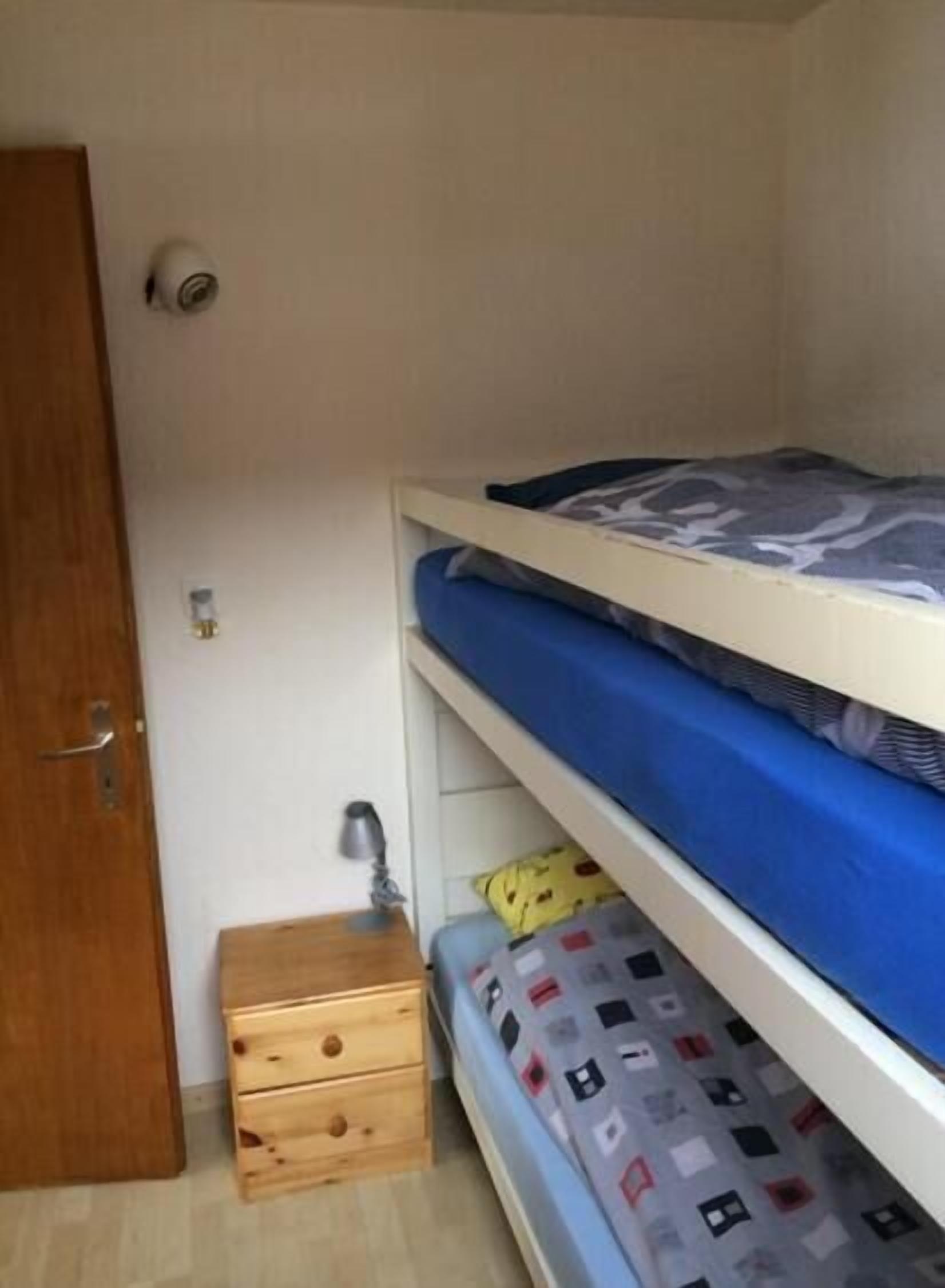 Una stanza piccola con un letto a castello, un mobiletto contenitore in legno, una porta e un punto luce a parete.