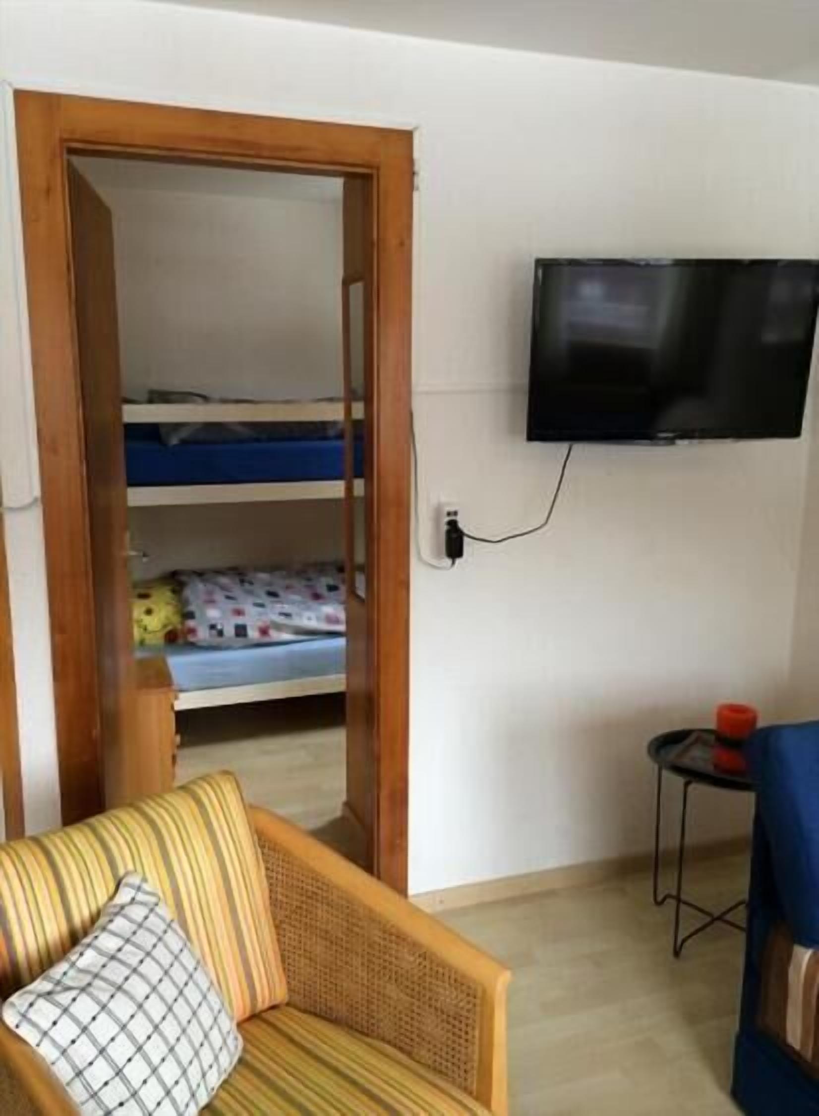 Une pièce avec un lit superposé, un téléviseur fixé au mur, une petite table sur laquelle se trouve un objet rouge, et un canapé à rayures.