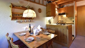 Une table de salle à manger en bois, mise avec des assiettes, des couverts et une bouteille de vin. Au-dessus de la table, on aperçoit un buffet en bois, et un coin cuisine avec un évier et une cuisinière.