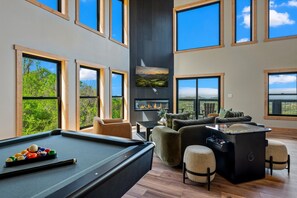 Ein modernes Wohnzimmer mit Billardtisch, bequemen Sitzmöglichkeiten, Kamin und großen Fenstern mit herrlichem Ausblick.
