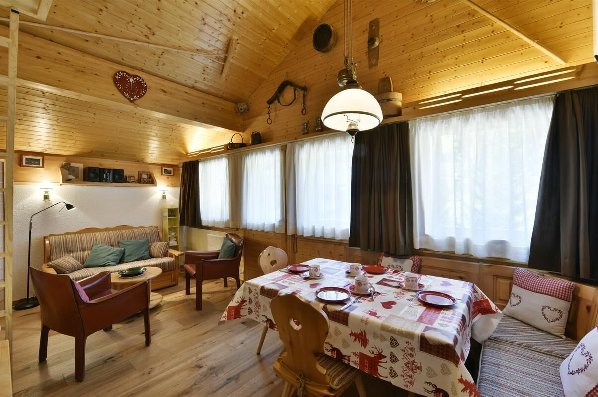A cozy cabin interior with a dining table set for four, wooden walls, a sofa, and a ceiling with a heart-shaped decoration.