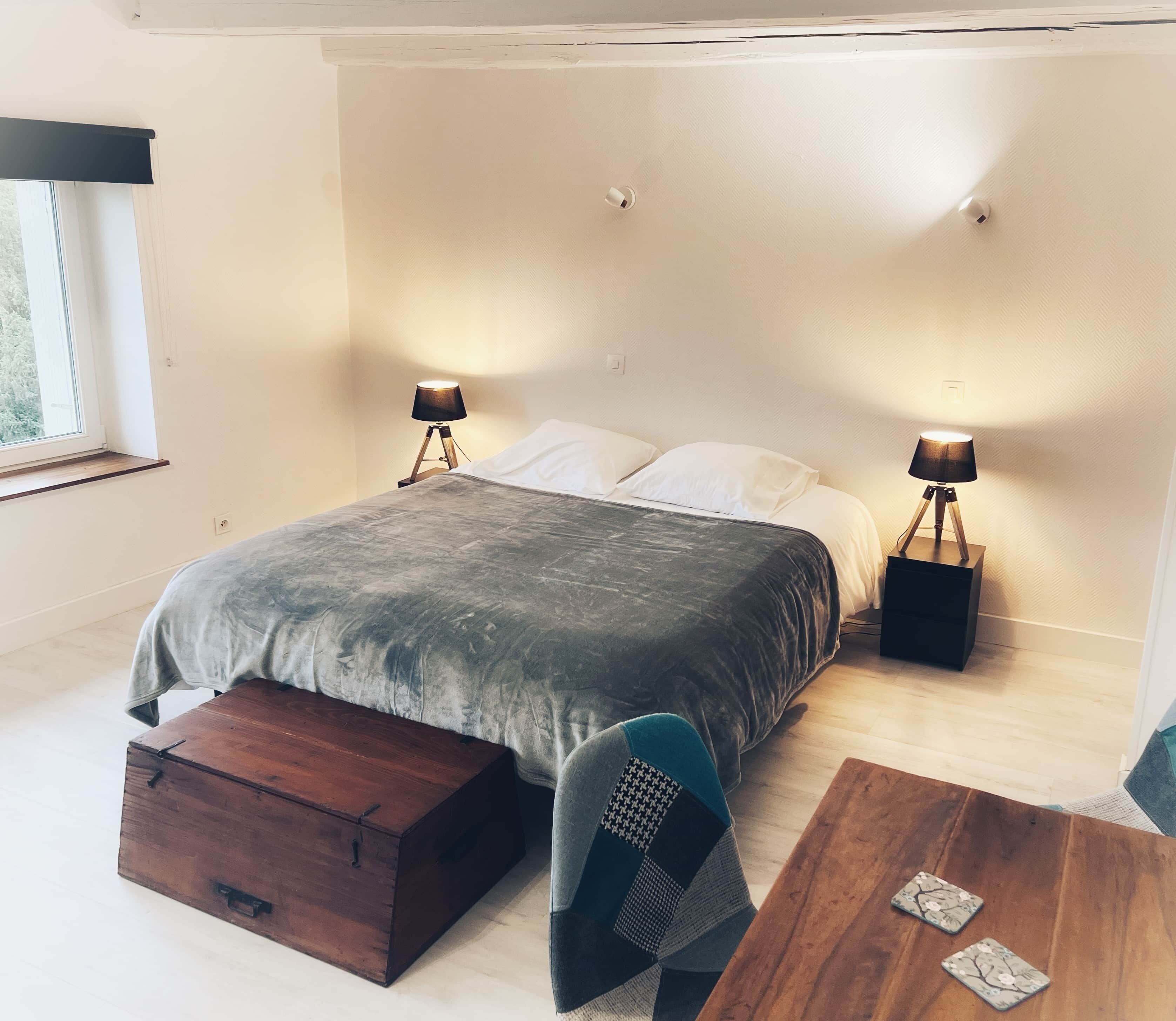 A modern bedroom with a large bed, two bedside lamps, a wooden chest, a small table, and a chair.