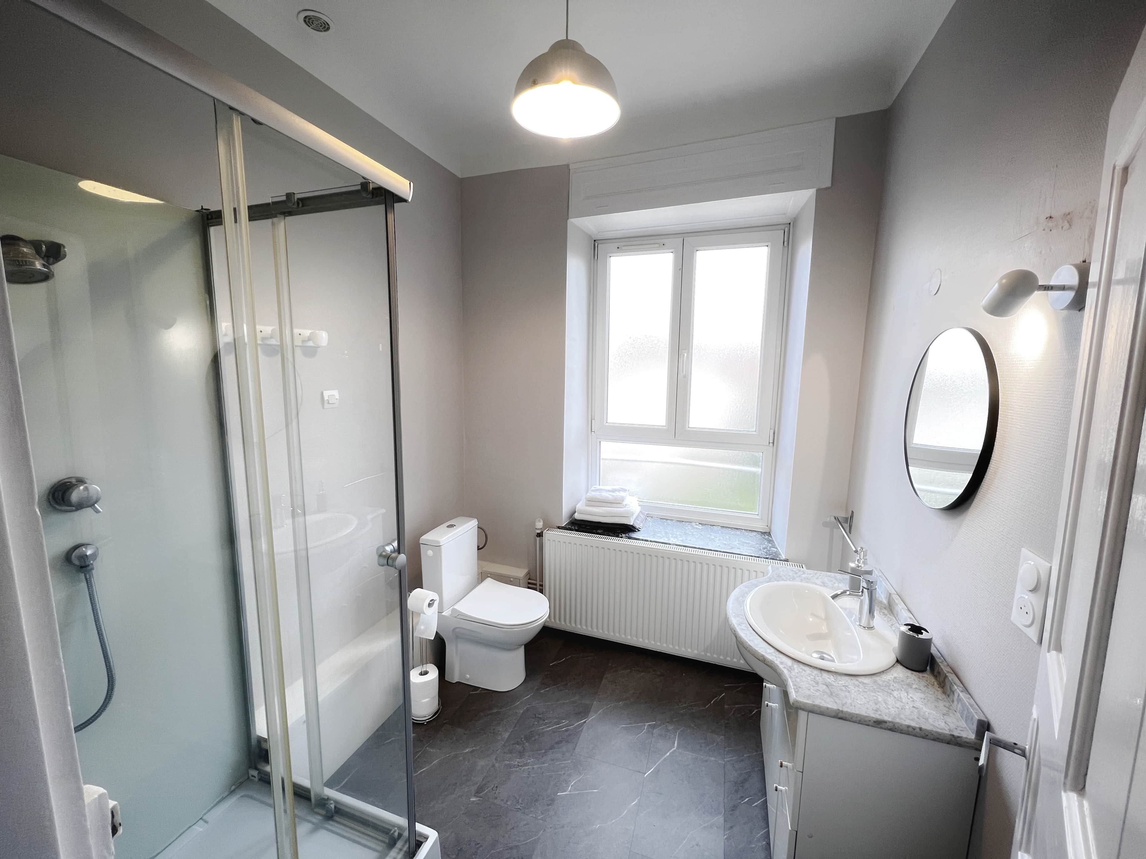 A modern bathroom with a glass shower enclosure, a toilet, a sink, and a window providing natural light.