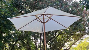 Eine Terrasse mit einem weißen Metalltisch und -stühlen, einem großen Sonnenschirm und einem Baum im Hintergrund.