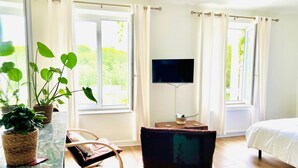 A hotel room with a bed, a TV mounted on the wall, a small table with a basket, a rocking chair, and large windows with curtains. - Studio 3, baignoire (Rurange-lès-Thionville)