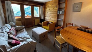 A cozy room with a wooden interior, a sofa, a small table, a chair, and a window with a view of mountains. - Comfortable 2  1/2-room apartment for 4 persons : living room - 1 alcove with 2 bunk beds - 1 room w (Grimentz)