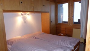 A wooden cabin-style bedroom with a bed, bedside tables, and a window with curtains. - Comfortable 2  1/2-room apartment for 4 persons : living room - 1 alcove with 2 bunk beds - 1 room w (Grimentz)