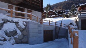 Un espacio exterior cubierto de nieve, con valla de madera, muro de piedra y un edificio con varias ventanas y puerta de madera.