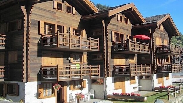 Una cabaña de madera con varios balcones, un cimiento de piedra y un paisaje montañoso de fondo.