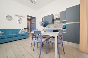 Ein modernes Wohnzimmer mit einem blauen Sofa, einem Esstisch mit Stühlen, einer Küchenzeile mit Schränken und einem blauen Gerät.