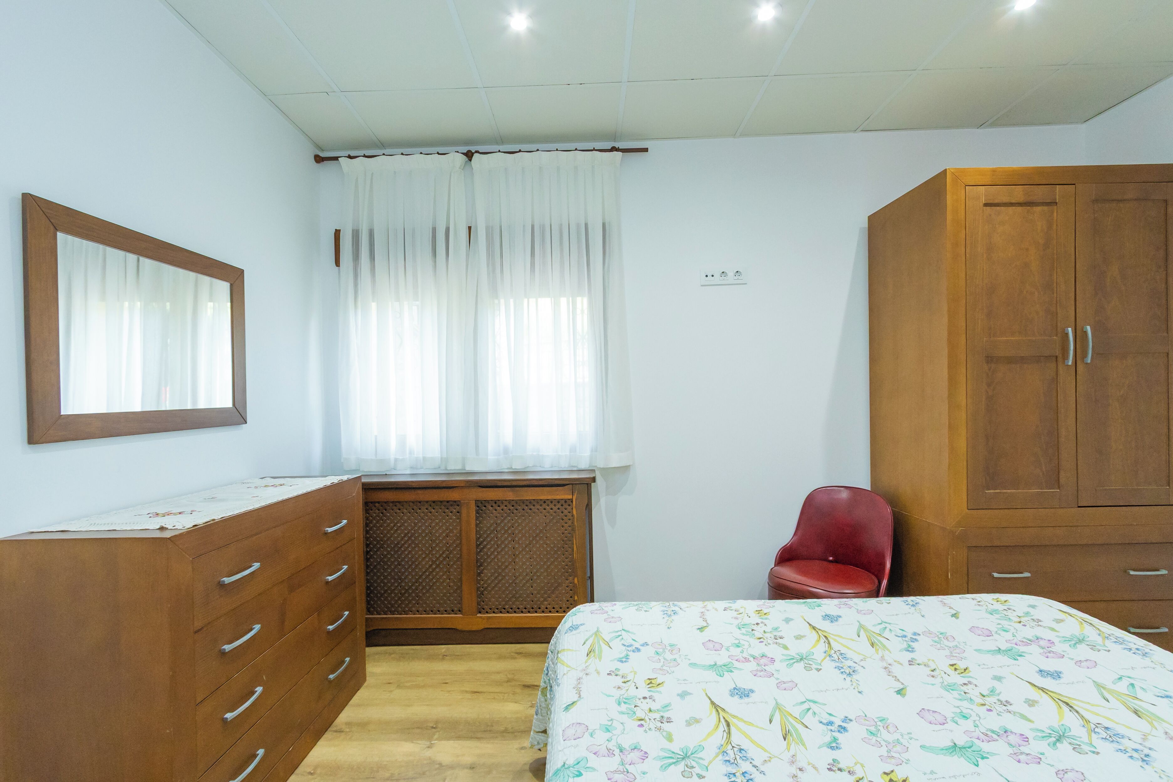 Een slaapkamer met een houten dressoir, een bed met bloemenbedekking, een rode stoel en een raam met dunne gordijnen.