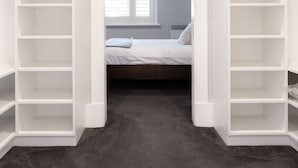 Ein begehbarer Kleiderschrank mit weißen Regalen, ein Bett mit weißer Bettwäsche und ein Fenster mit Jalousien.