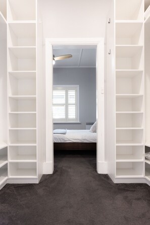 Ein begehbarer Kleiderschrank mit weißen Regalen, ein Bett mit weißer Bettwäsche und ein Fenster mit Jalousien.
